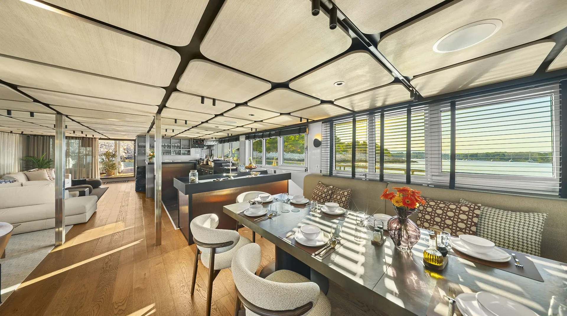 Modern yacht interior with a dining table set for six, an open kitchen, large windows with blinds, sunlight streaming in, and scenic water views outside. Contemporary furniture and decor create a bright, inviting space.
