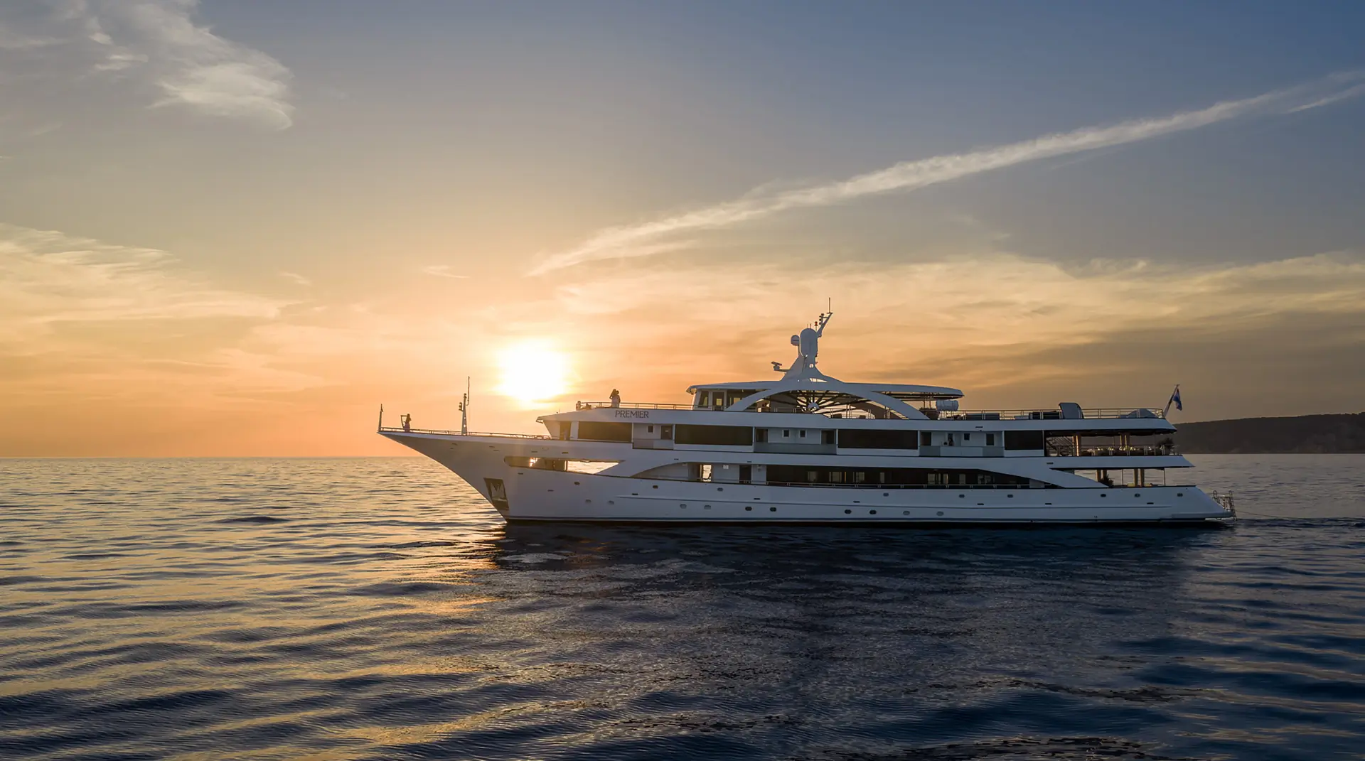 A large white yacht sails on calm water at sunset, with the golden sun low in the sky and a distant coastline visible on the horizon.