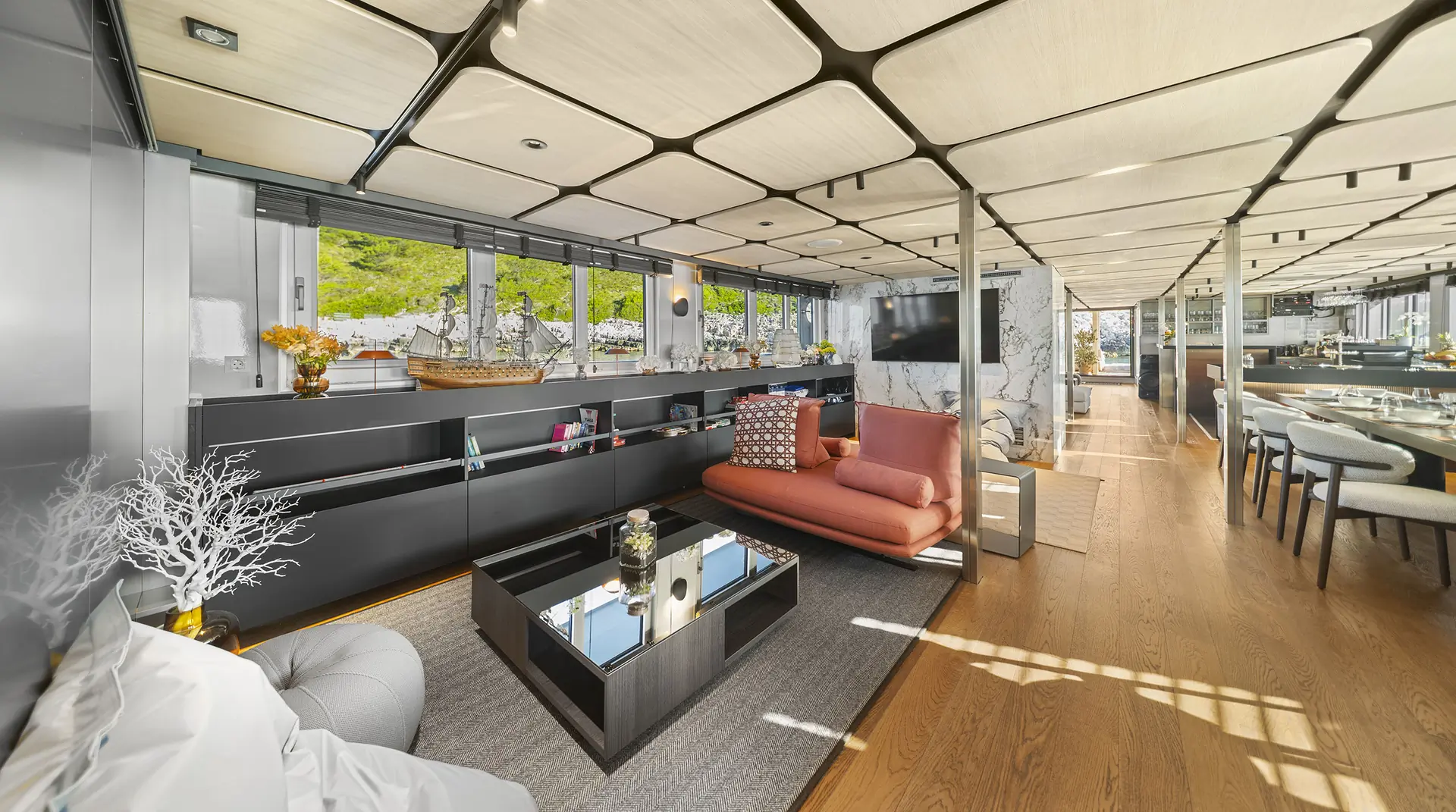 Modern, open-plan living area with wood floors, large windows, and contemporary furniture, including a pink sofa and glass coffee table. Natural light fills the space, and green hills are visible outside.