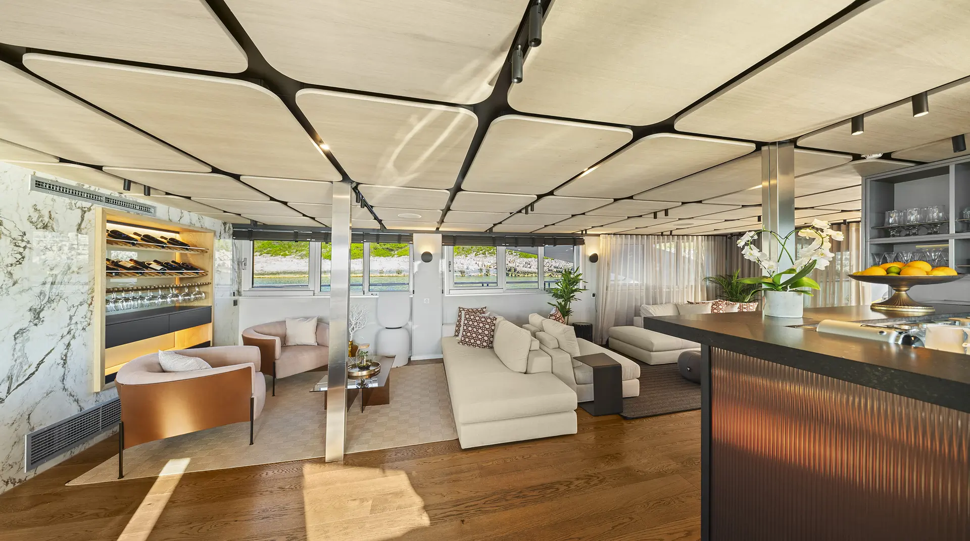 Luxurious yacht lounge with modern white sofas, armchairs, and a marble wine rack. Large windows offer scenic views, and the wooden floor and geometric ceiling add to the elegant ambiance.