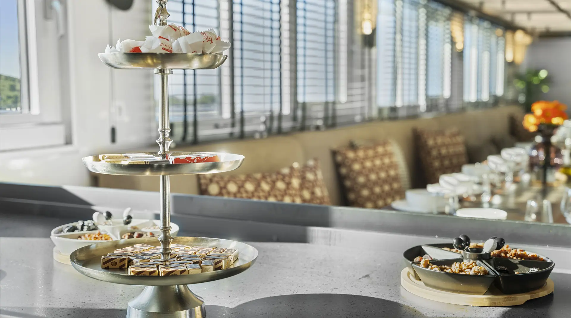 A three-tiered metal tray holds assorted pastries and sweets on a countertop in a bright, modern dining area with tables and chairs in the background.