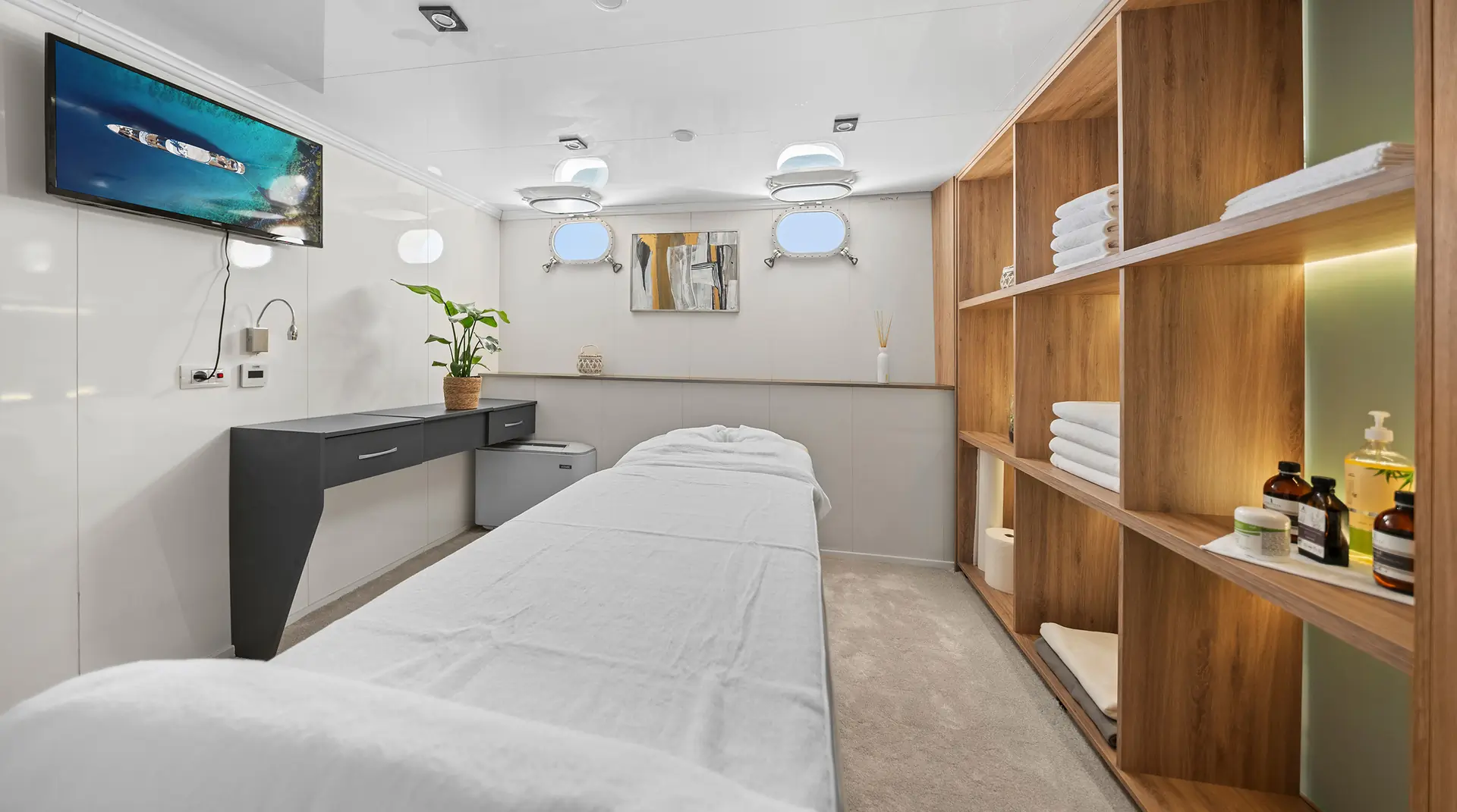 A modern spa treatment room with a massage table covered in white linens, wooden shelves storing towels and oils, a potted plant, a wall-mounted TV, and small windows providing natural light.