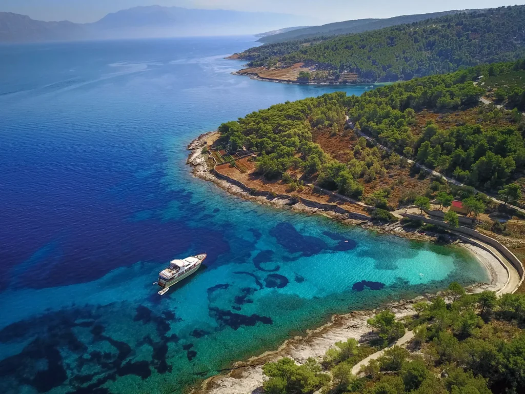 KORAB Aerial view of a clear blue sea with a boat near a rocky coastline covered in green trees and vegetation, with hills and distant land visible on the horizon.