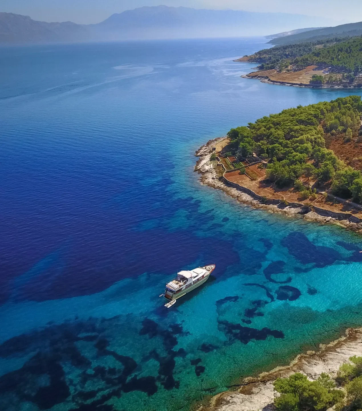 KORAB Aerial view of a yacht anchored near a rocky, forested coastline with clear turquoise water. The lush green peninsula extends into the sea, and distant hills are visible under a clear blue sky.