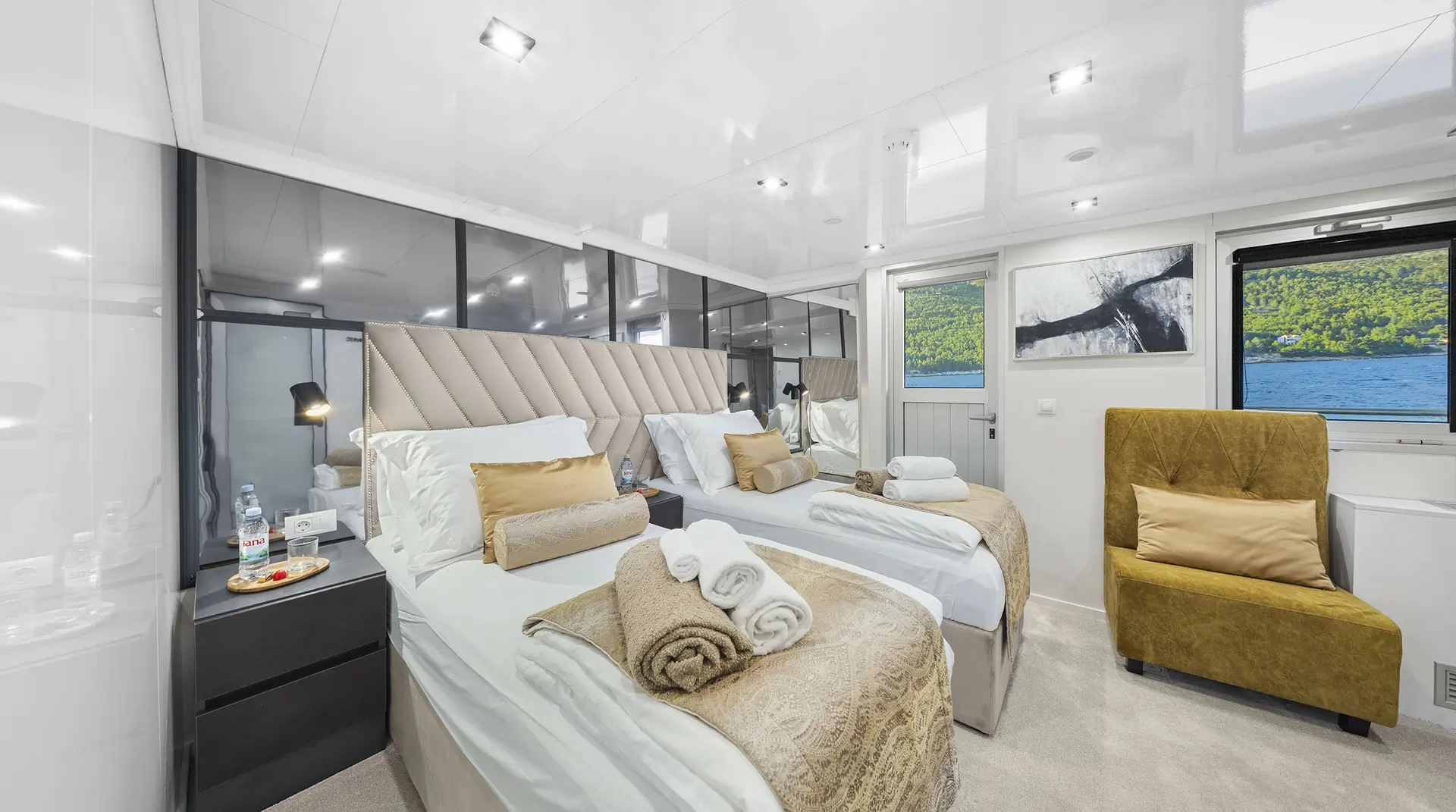 Modern, bright cruise ship cabin with two twin beds, gold and beige bedding, a towel arrangement, a green armchair, nightstand with water and fruit, large wall mirror, art on the wall, and a window with a view of water and greenery.