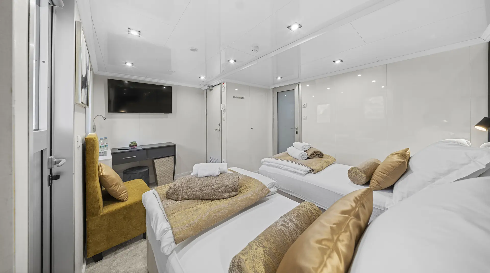 Modern, well-lit cabin with two single beds, gold accent pillows, a mustard yellow chair, a flat-screen TV, and a small desk with water bottles; white walls and bedding create a clean, cozy atmosphere.