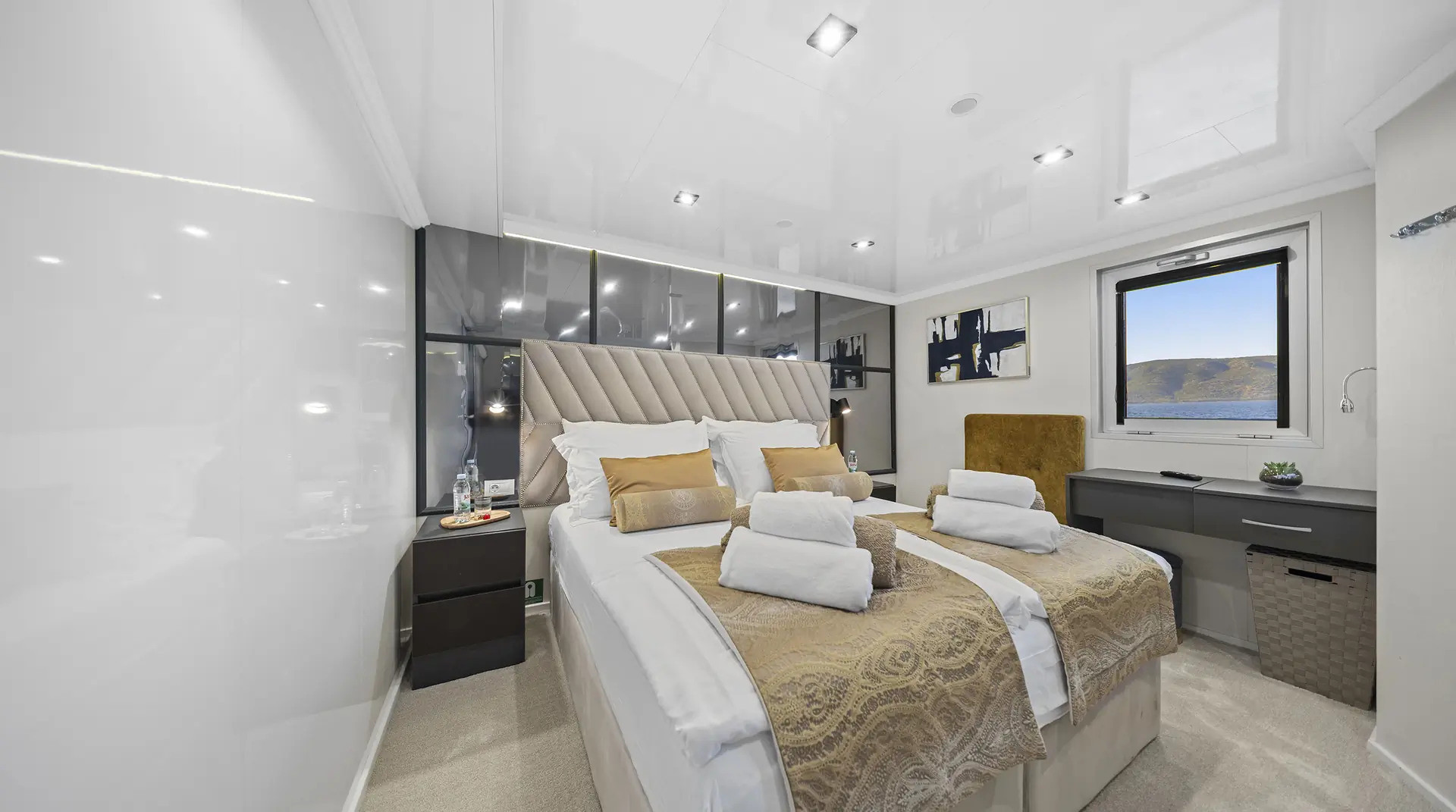 A modern, well-lit bedroom with a large bed dressed in gold and white linens, two bedside tables, towel sets, a desk by a window with a scenic view, and contemporary decor on the walls.