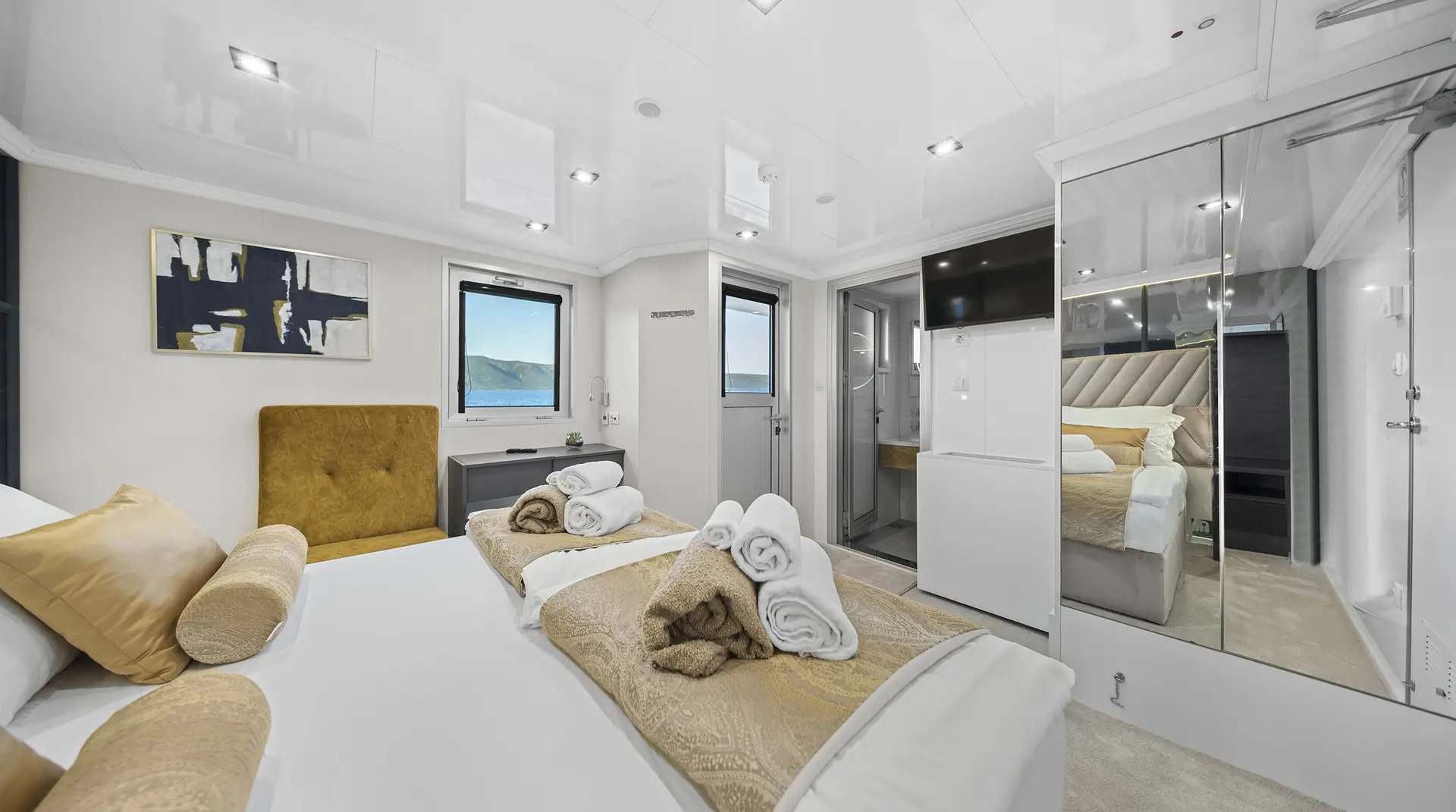 A modern, bright bedroom with two single beds, beige and gold bedding, rolled towels, a mustard headboard, wall art, a small desk, mirrored closet, and an en-suite bathroom. A window shows mountain scenery outside.