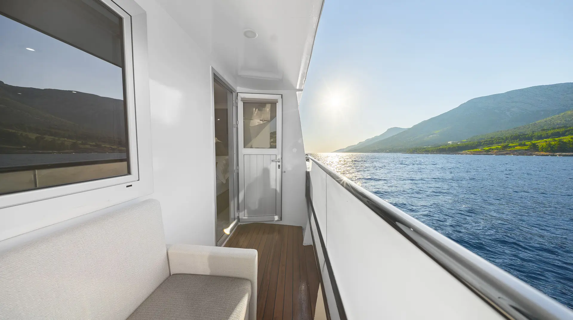 A view from a yacht balcony with a white sofa, wooden floor, and glass door, overlooking a calm blue sea and distant green mountains under a bright, clear sky.