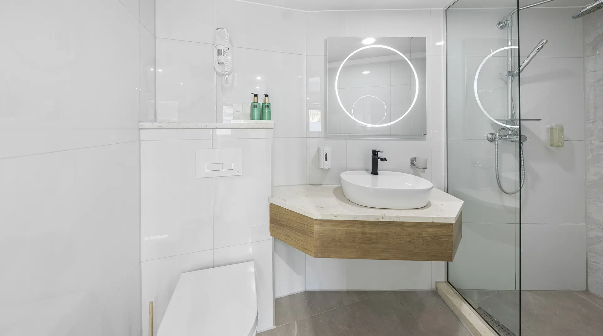 Modern bathroom with white tile walls, a glass shower enclosure, a round illuminated mirror above a vessel sink on a wood-accented counter, and a wall-mounted toilet. Three green soap bottles are on a shelf.