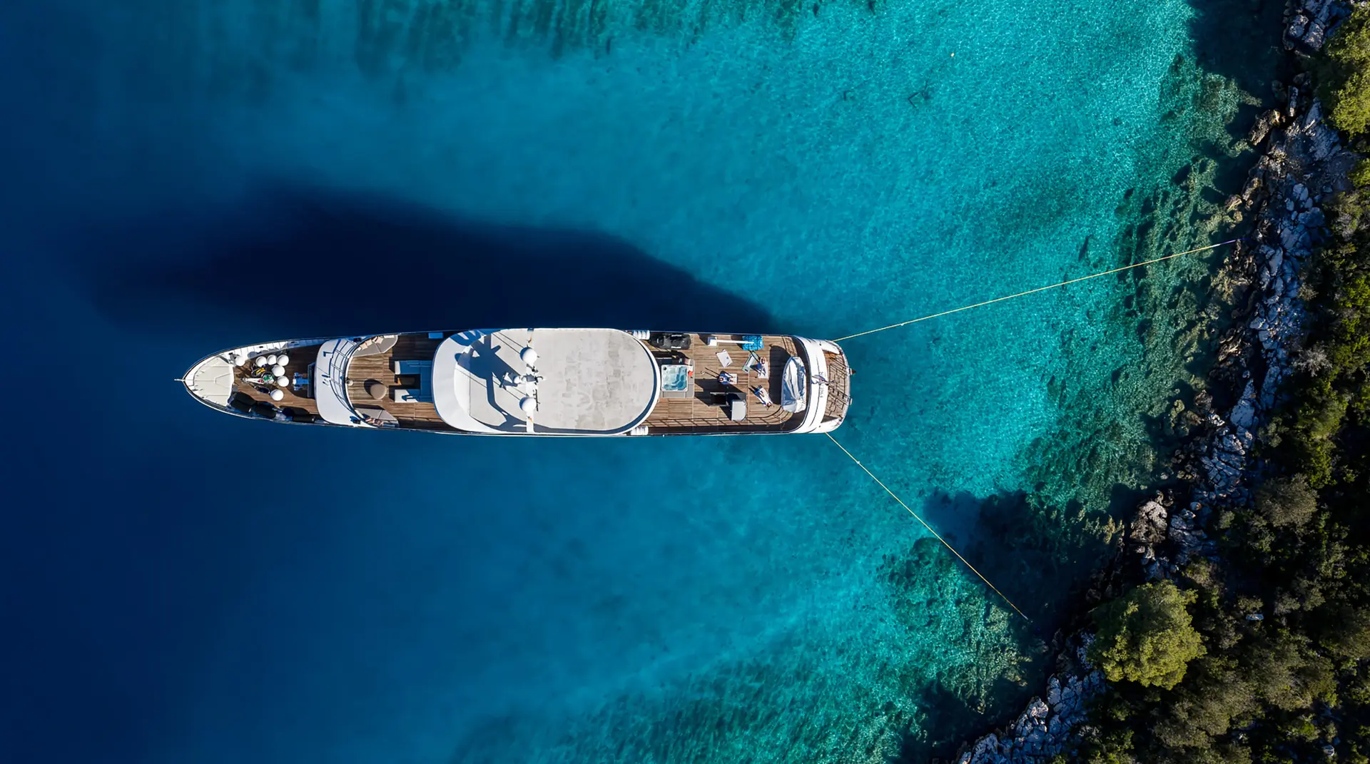 Aerial view of a large yacht anchored in clear turquoise water near a rocky, tree-lined shoreline, with two ropes securing the boat to the land.