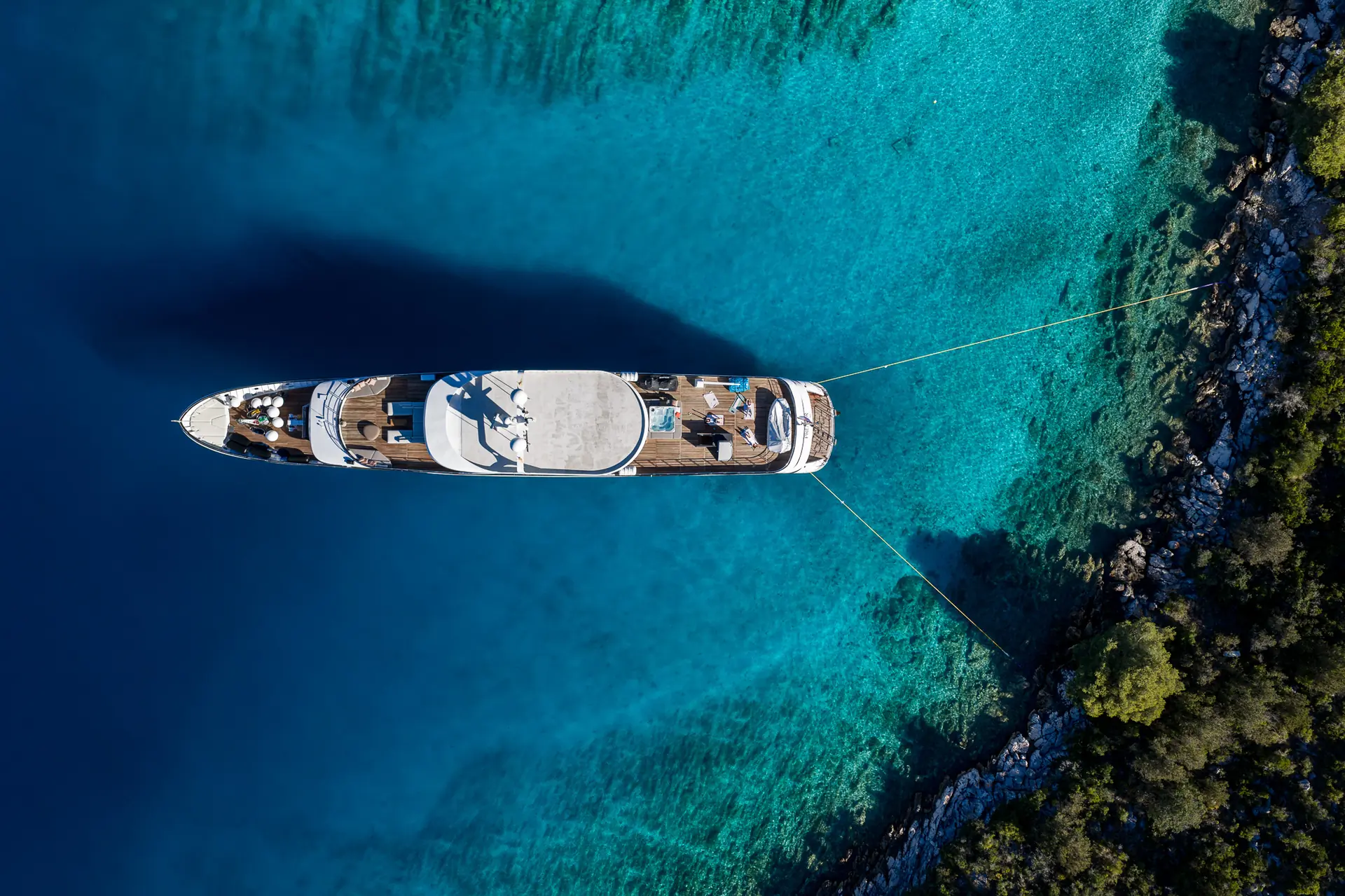 Aerial view of a large white yacht anchored in clear turquoise water near a rocky, tree-lined shore. Two ropes secure the yacht to the land, and sunbathers can be seen on the deck.