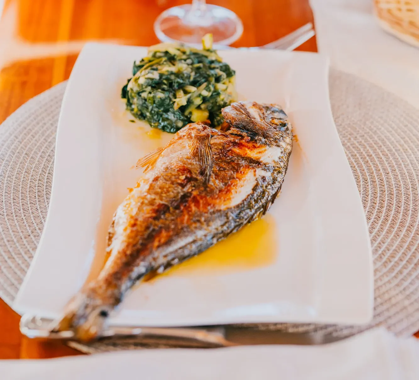 A grilled whole fish served on a white plate with a side of sautéed greens, accompanied by a glass of white wine on a wooden table.
