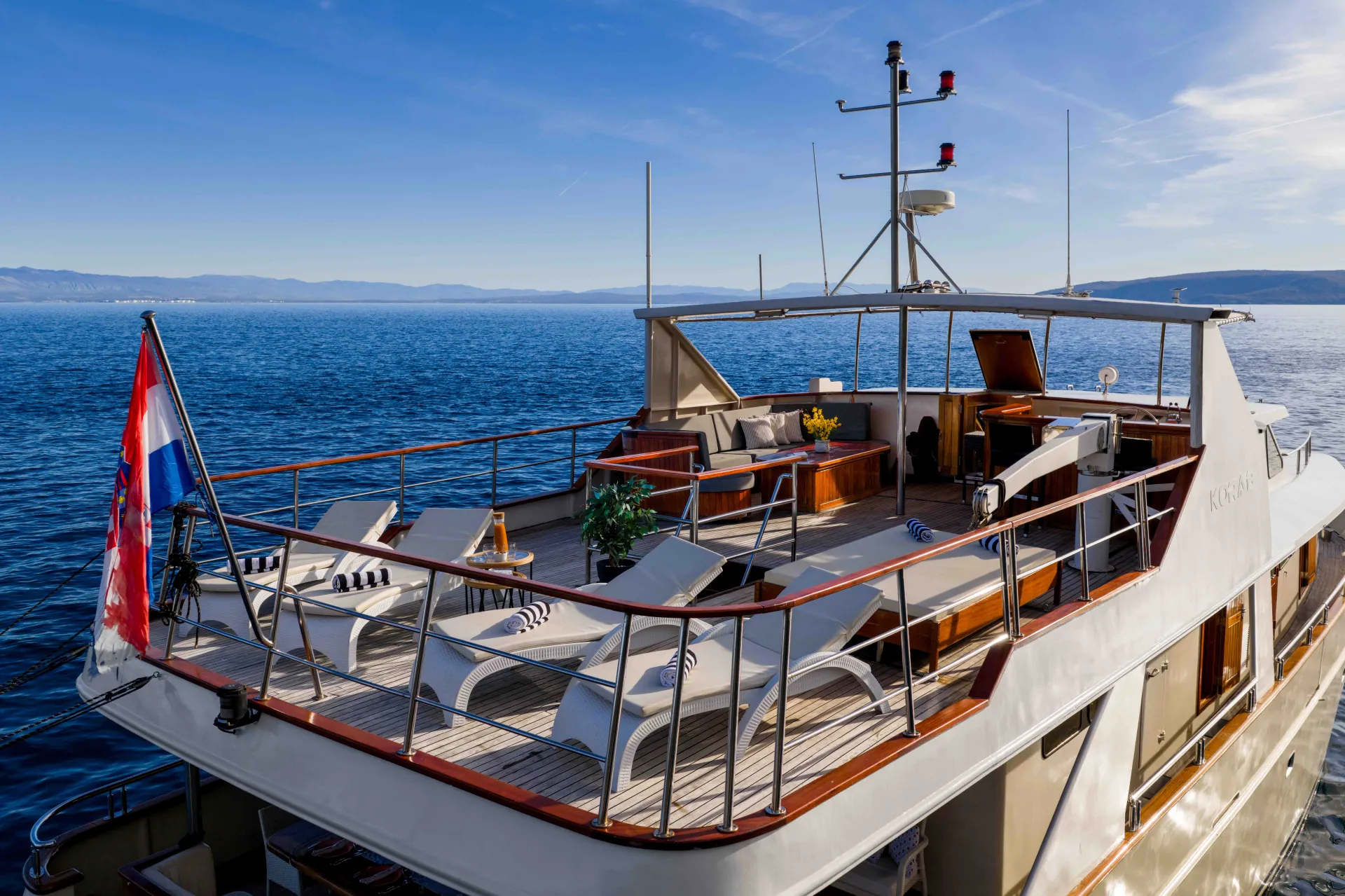KORAB A luxury yacht deck with lounge chairs, tables, and plants overlooks calm blue water under a clear sky. A flag is displayed at the back, and distant land is visible on the horizon.