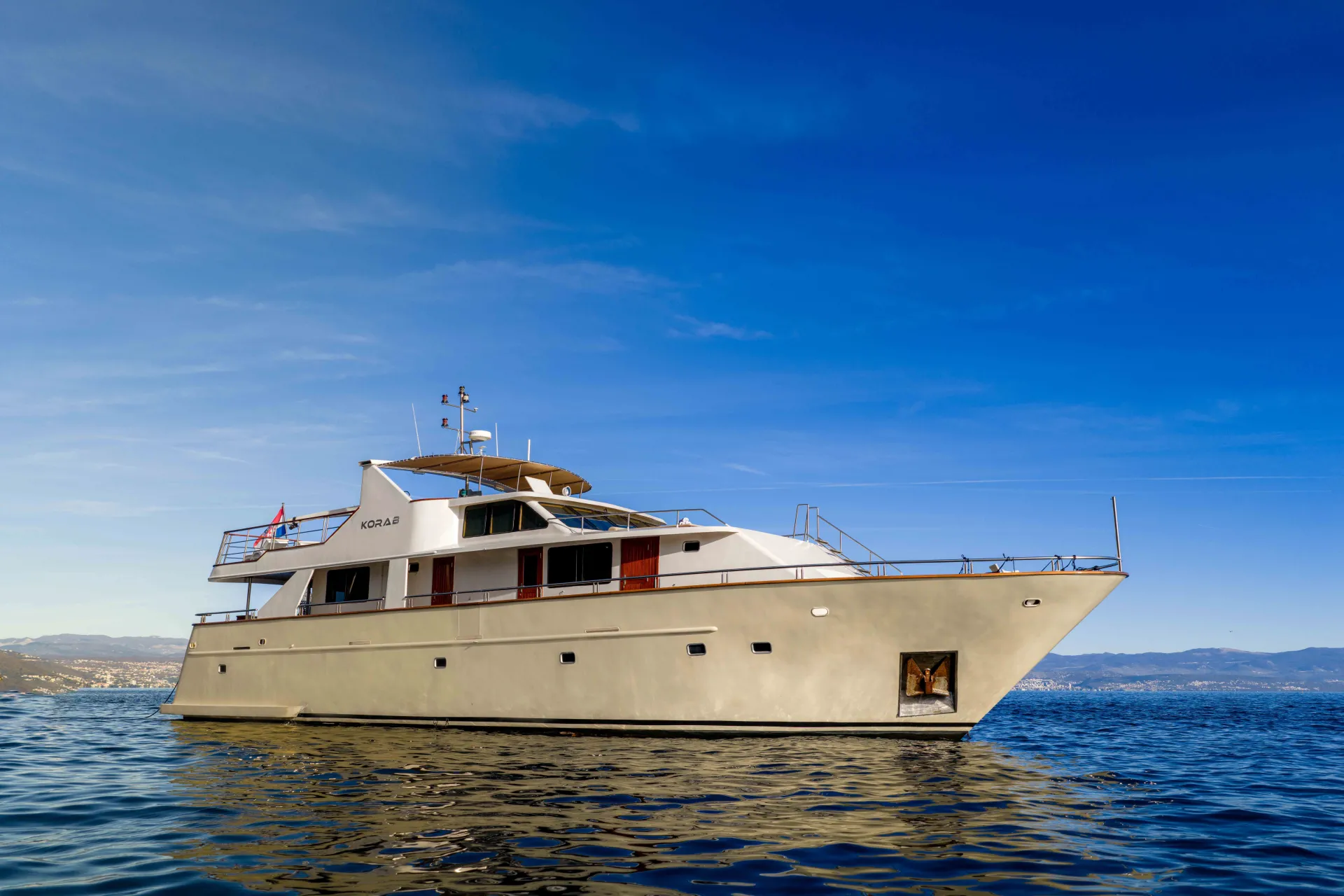 KORAB A white motor yacht floats on calm blue water under a clear sky, with distant hills and a coastal city visible in the background.