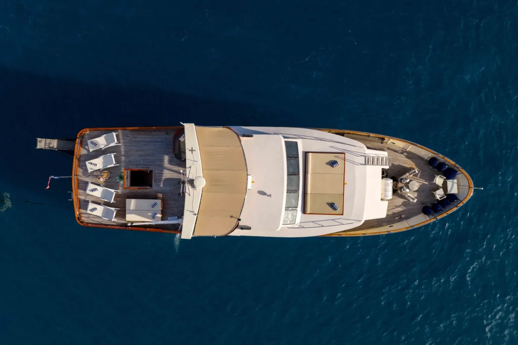 KORAB Aerial view of a luxury yacht on clear blue water, showing deck areas with seating, tables, and sun loungers. The yacht is stationary, and its shadow is visible in the water below.