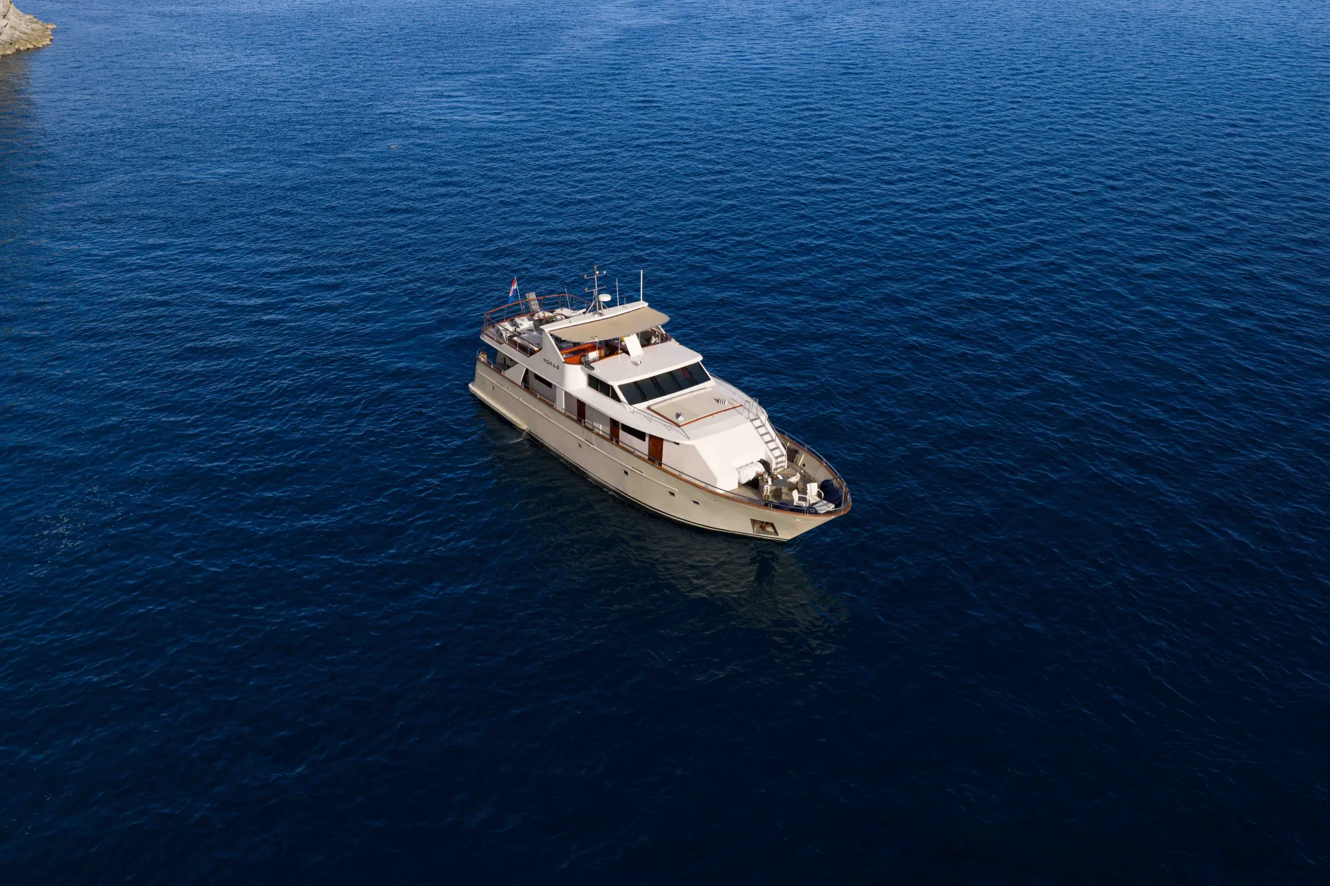 KORAB A white motor yacht floats alone on deep blue, calm ocean water under clear skies, viewed from above.