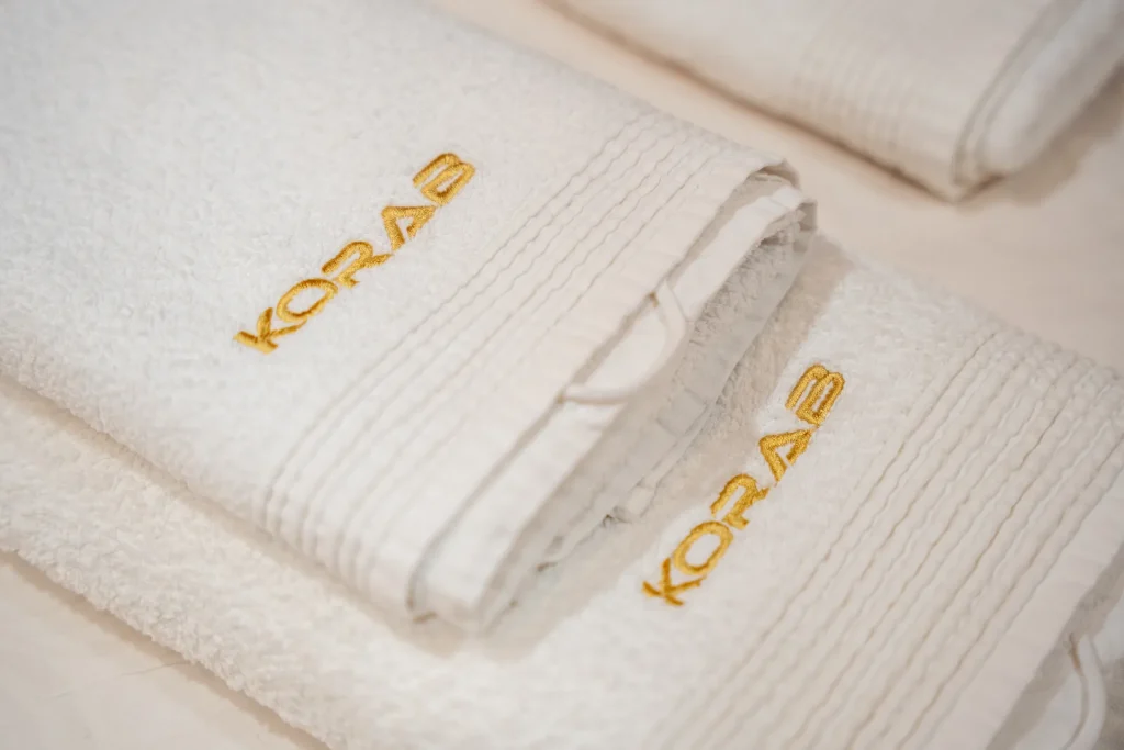 KORAB Two neatly folded white towels with KORAB embroidered in gold thread are stacked on a white surface. Another similar towel is partially visible in the background.