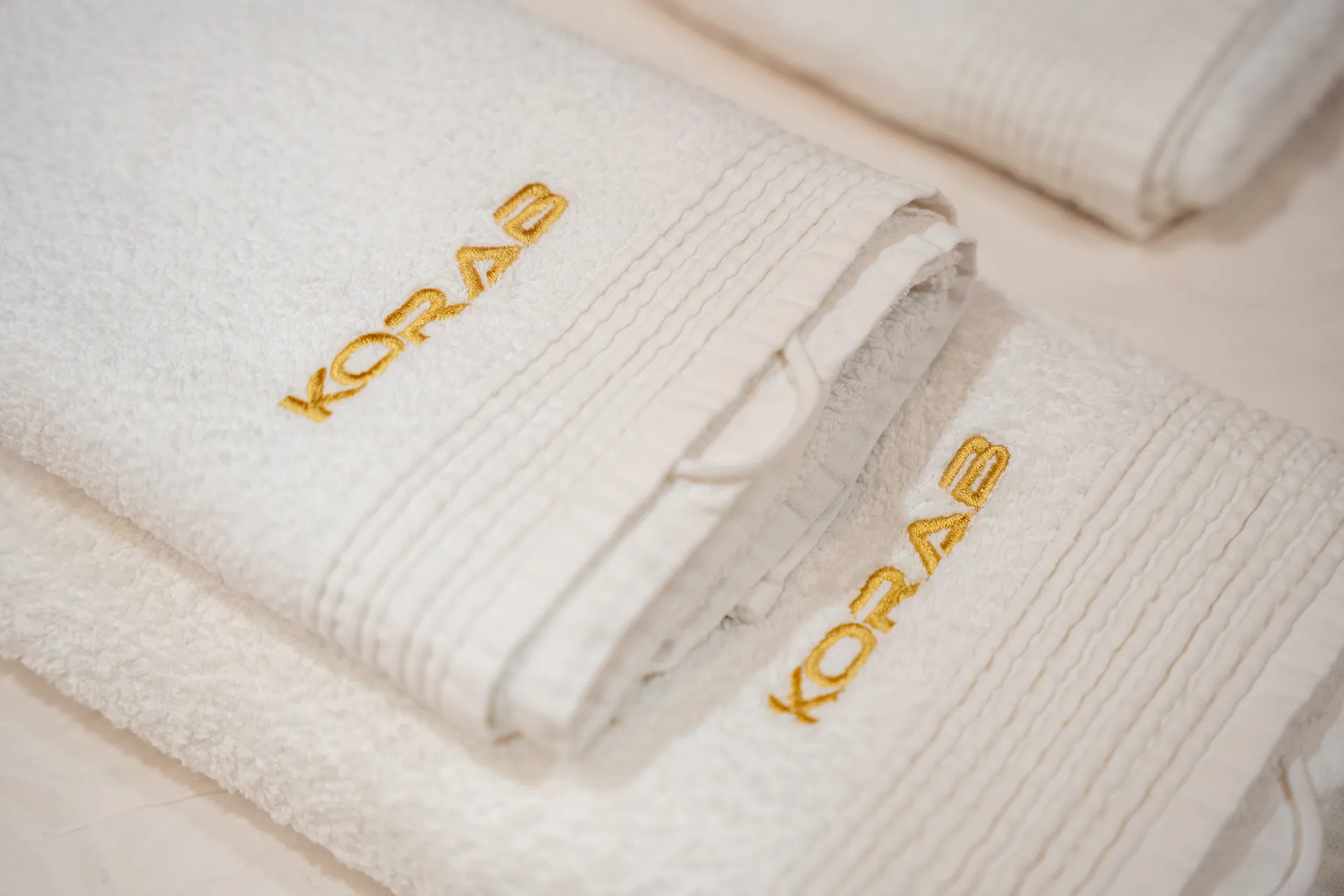 KORAB Two neatly folded white towels with KORAB embroidered in gold thread are stacked on a white surface. Another similar towel is partially visible in the background.