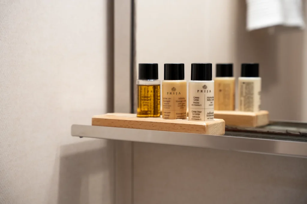 KORAB A close-up of several small bottles of toiletries, including shampoo and shower gel, arranged neatly on a wooden tray on a bathroom shelf. A mirror in the background reflects the bottles.