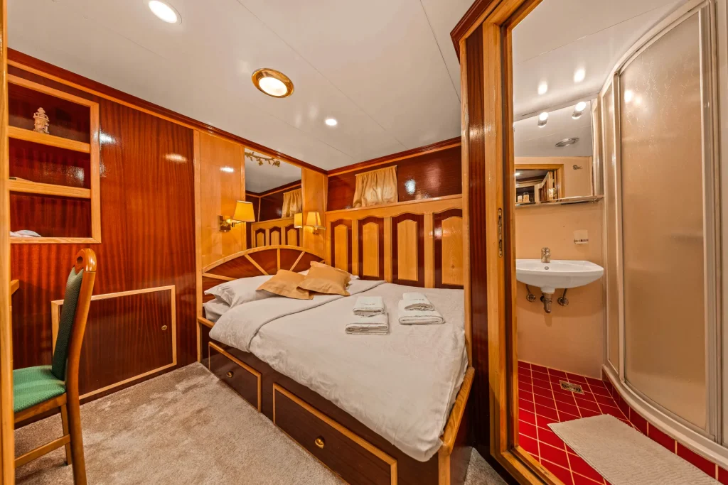 KORAB A cozy cabin room with wood-paneled walls, a double bed with folded towels, built-in shelves, a green chair, and an adjoining bathroom featuring a sink and a corner shower with a red-tiled floor.