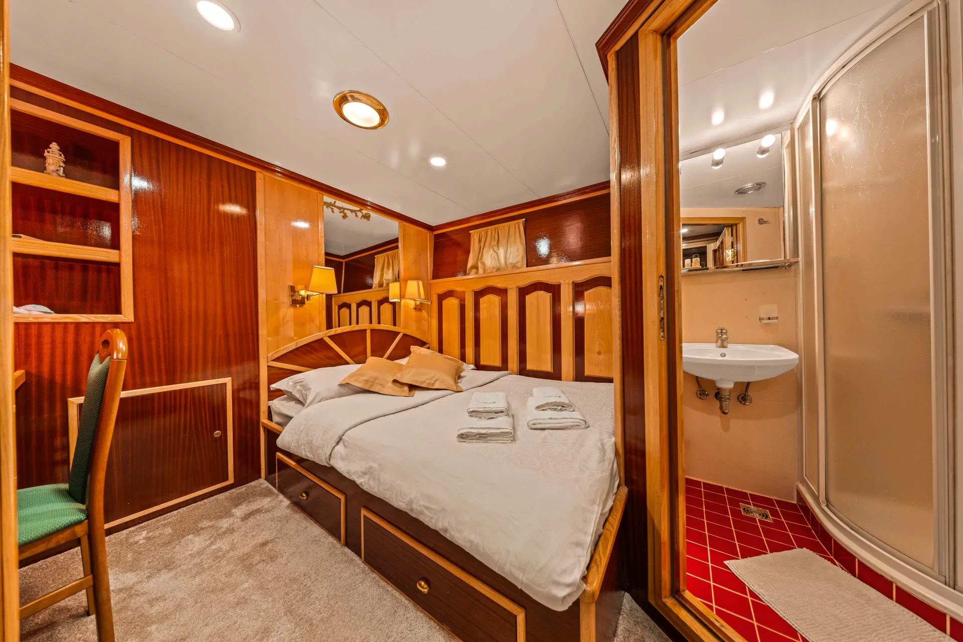 KORAB A cozy cabin room with wood-paneled walls, a double bed with folded towels, built-in shelves, a green chair, and an adjoining bathroom featuring a sink and a corner shower with a red-tiled floor.