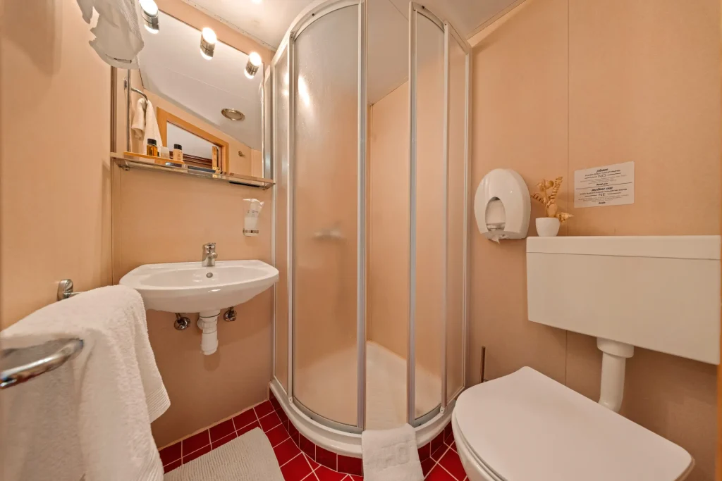 KORAB A compact bathroom with a red tiled floor, white sink, round shower enclosure with sliding doors, toilet, mirror, towel rack, and a soap dispenser on the wall.