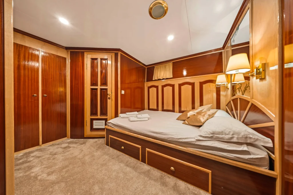 KORAB Cozy bedroom with polished wood paneling, a double bed with white linens and beige pillows, two folded towels on the bed, built-in closets, soft beige carpet, and warm ambient lighting.