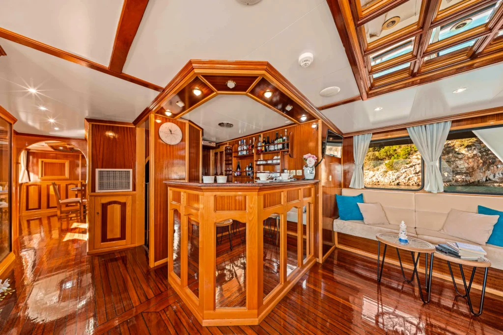 KORAB Luxurious yacht interior with polished wooden floors, a central bar area with shelves of bottles, a clock, cozy seating with blue pillows, side tables, and large windows with curtains showing a scenic outdoor view.