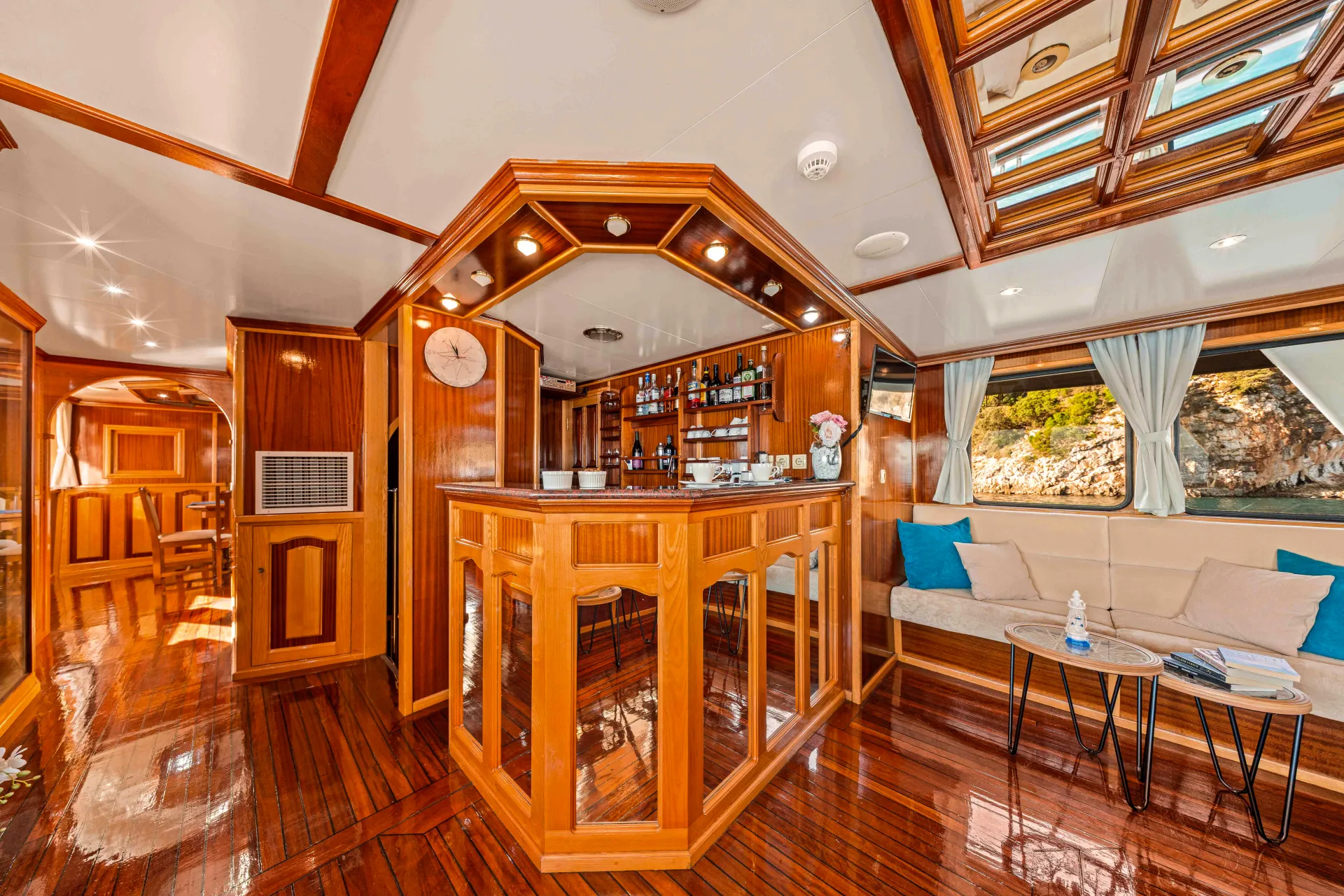 KORAB Luxurious yacht interior with polished wooden floors, a central bar area with shelves of bottles, a clock, cozy seating with blue pillows, side tables, and large windows with curtains showing a scenic outdoor view.