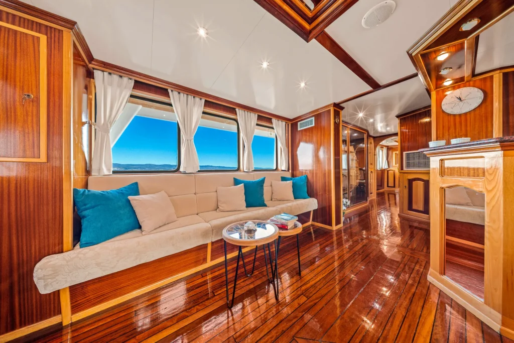 KORAB Luxurious yacht interior with polished wooden floors, cream-colored sofa with blue and beige pillows, round coffee tables, large windows with white curtains, and natural light streaming in. Ocean and sky visible through windows.