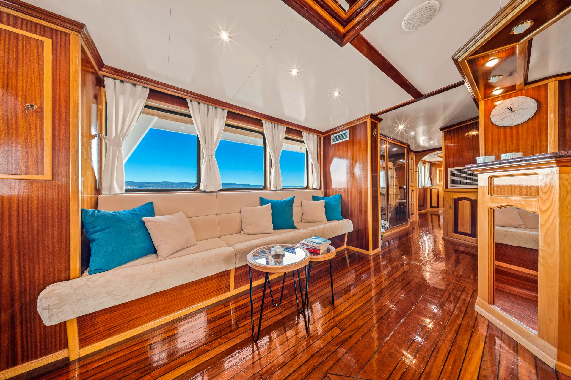 KORAB Luxurious yacht interior with polished wooden floors, cream-colored sofa with blue and beige pillows, round coffee tables, large windows with white curtains, and natural light streaming in. Ocean and sky visible through windows.