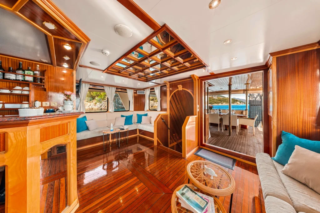 KORAB A luxurious yacht interior with polished wood floors, cream sofas with blue pillows, a bar area, and large windows. Glass doors open to an outdoor dining area with ocean views.