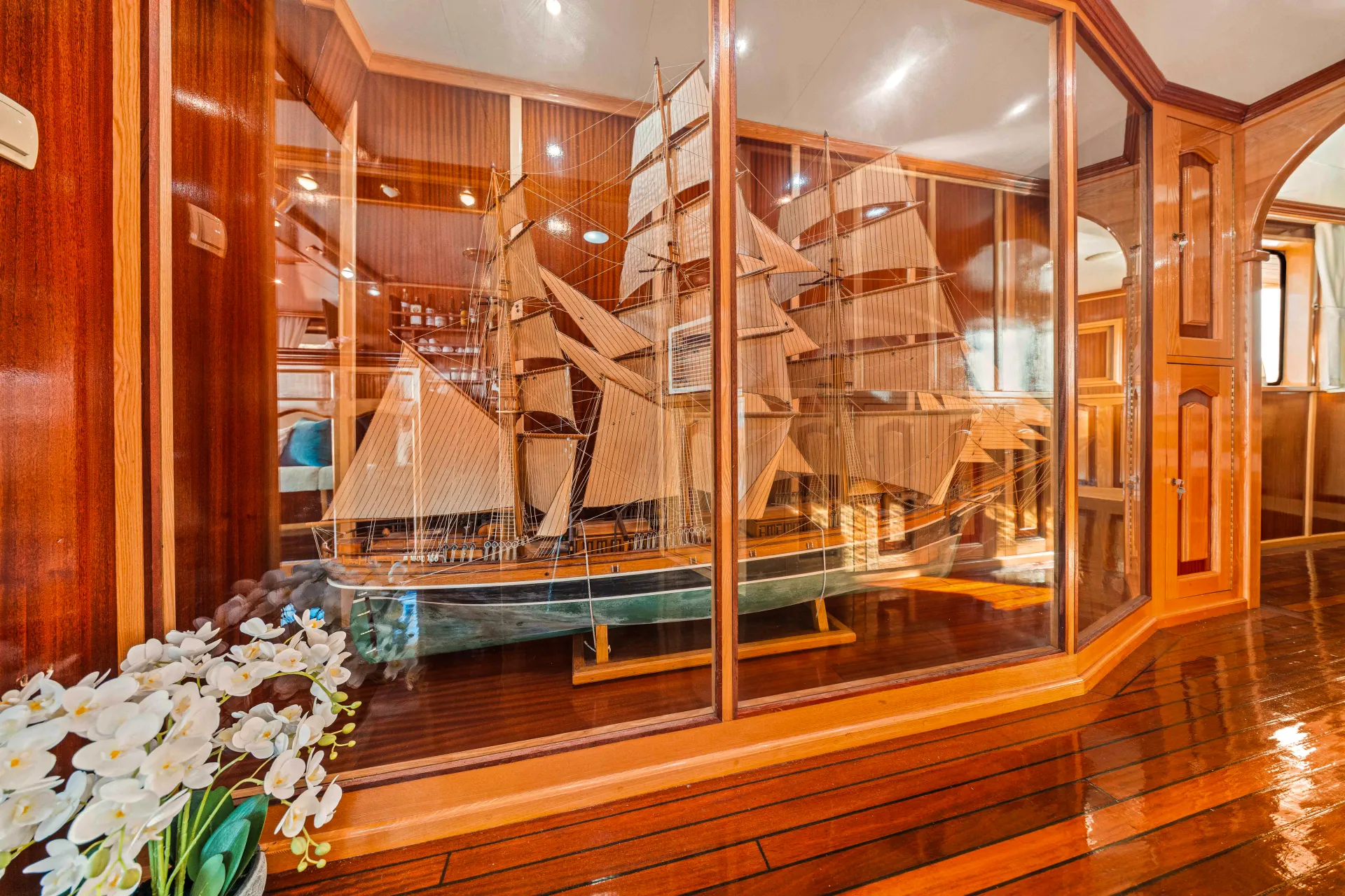 KORAB A detailed wooden model ship with multiple sails is displayed in a large glass case in a wood-paneled room. White flowers sit on the polished wooden floor in the foreground.