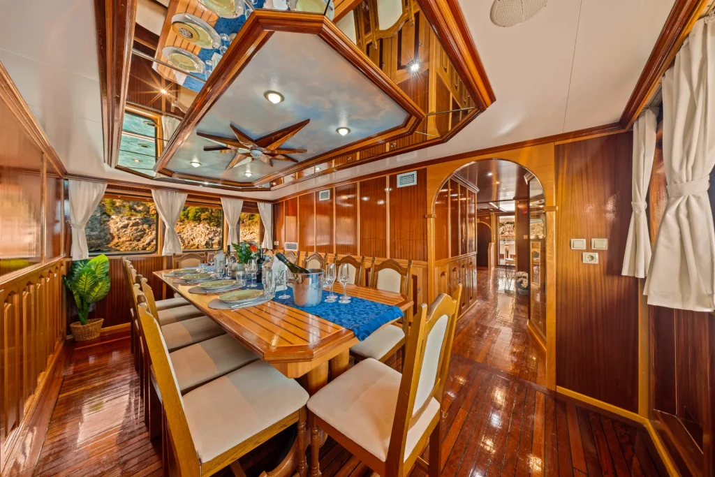 KORAB Luxurious yacht dining room with polished wood walls and floors, an elegant dining table set for eight, large windows with white curtains, and a decorative ceiling with a nautical theme.