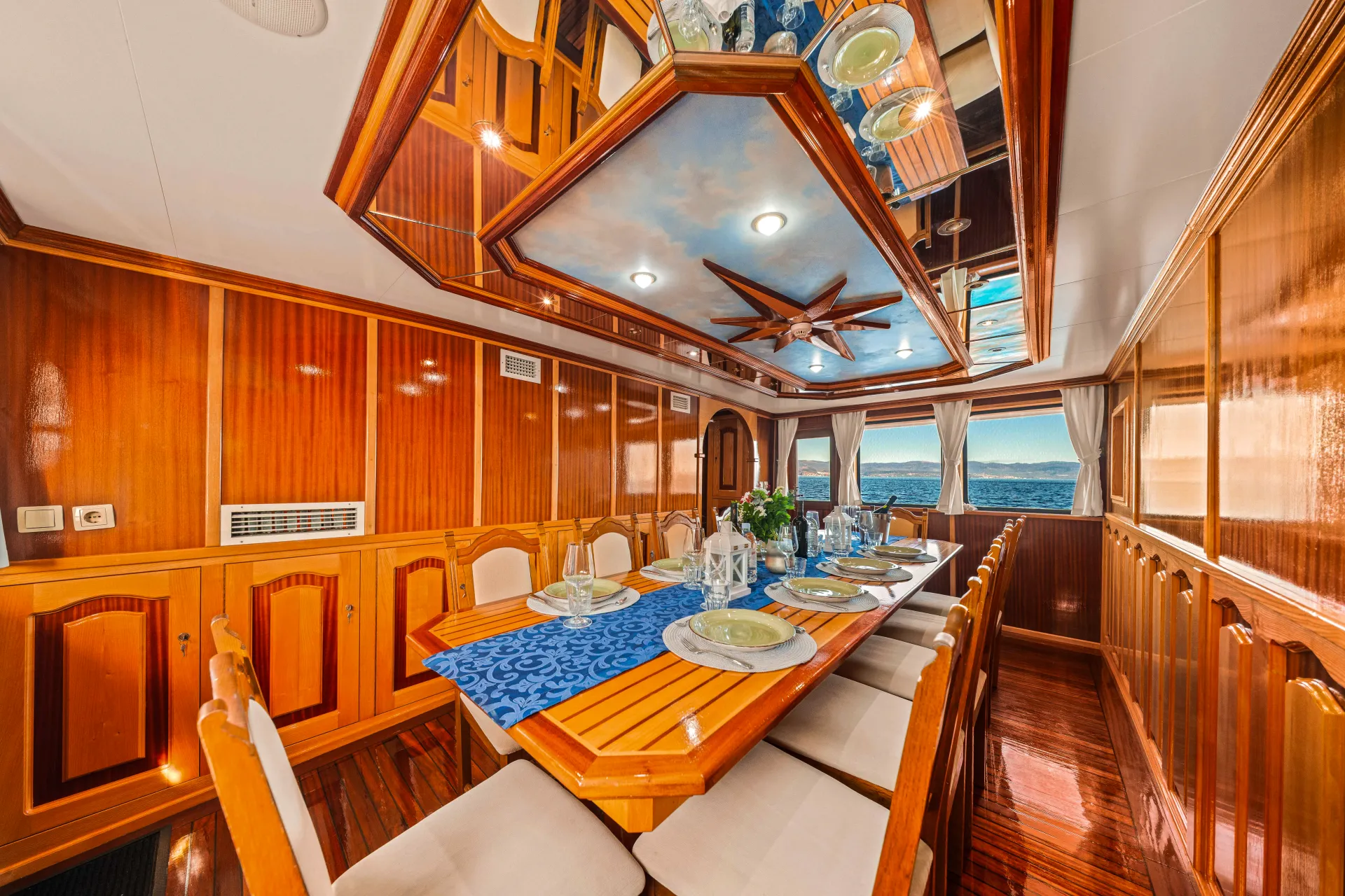 KORAB A luxurious yacht dining room featuring a wooden table set for eight, cushioned chairs, polished wood-paneled walls, large windows with ocean views, and a mirrored ceiling with sky and fan designs.