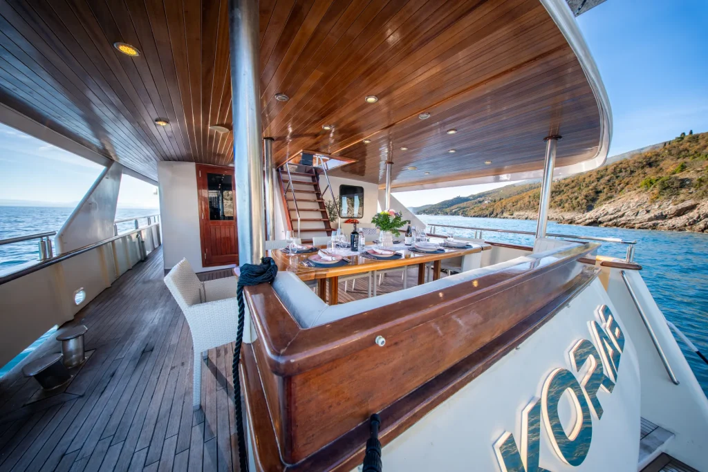 KORAB Luxurious yacht deck with a polished wooden ceiling, dining table set for a meal, cushioned seats, and ocean views; coastline visible in the background under a clear sky.