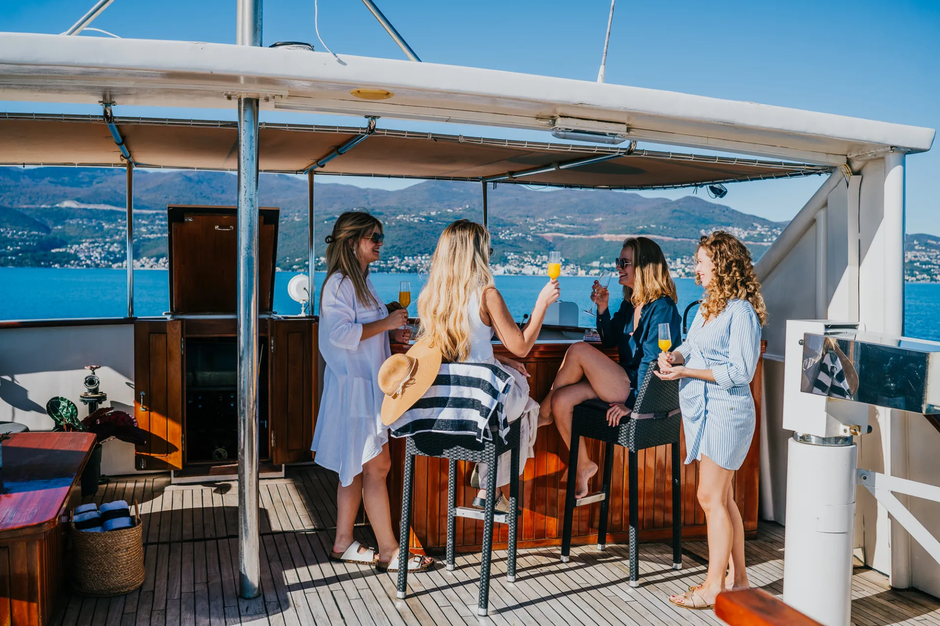 KORAB Four women sit and stand around an outdoor bar on a yacht, holding drinks and talking. The weather is sunny, with blue water and distant mountains visible in the background.