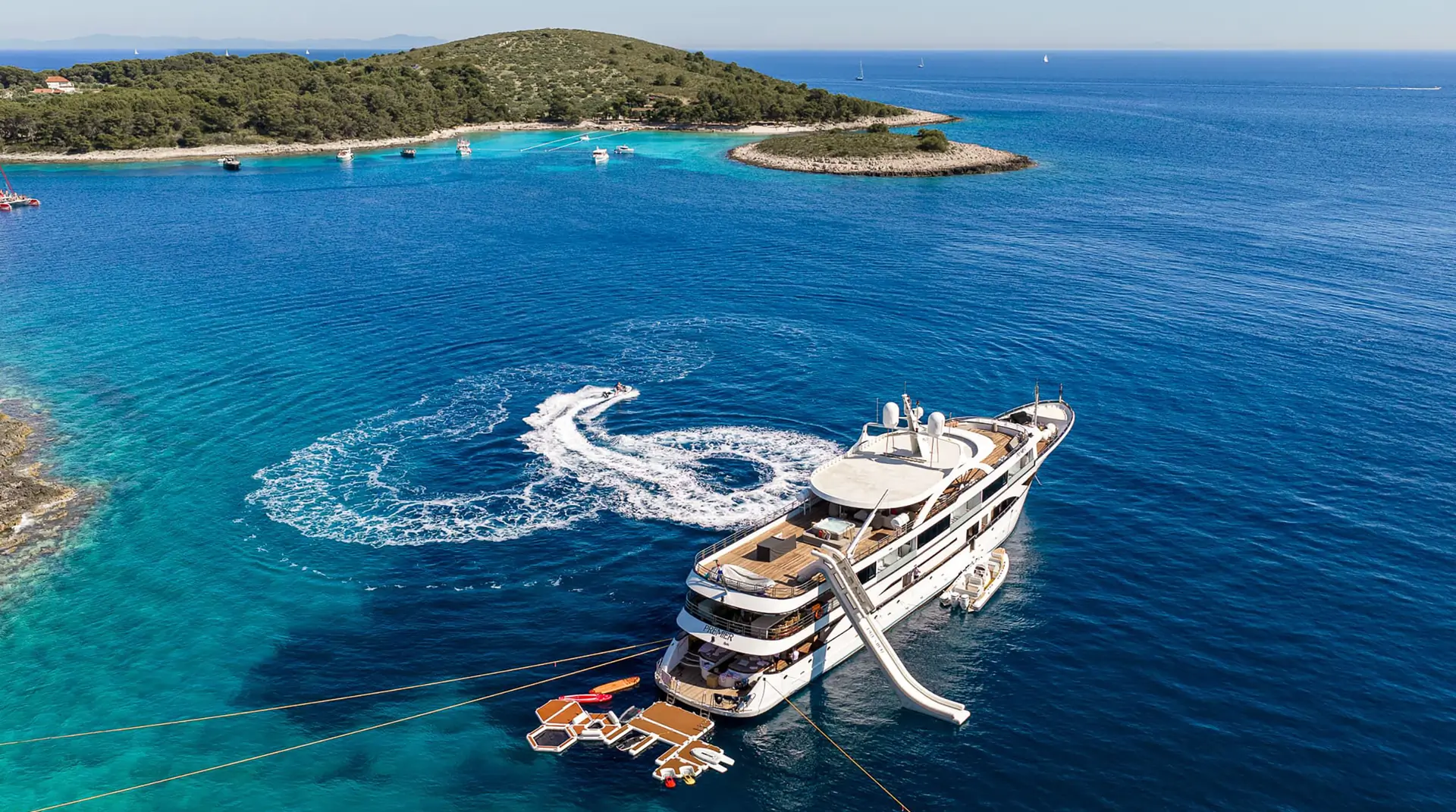 A luxury yacht anchored near a small island with clear blue water, water toys and a slide off the yacht, and a jet ski creating a swirl pattern in the sea.