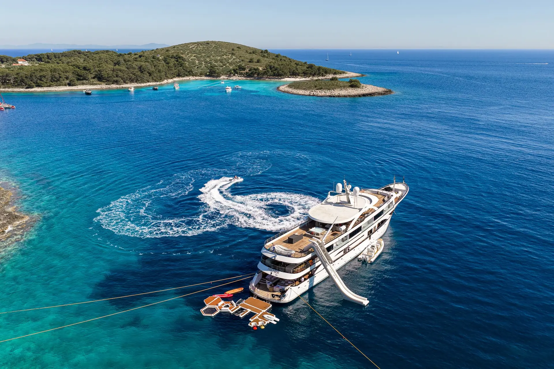 A large yacht is anchored in clear blue water near a small green island, with a slide extending from the yacht and a swirl pattern in the water behind a small boat. Floating docks and lounge chairs are attached to the yacht.