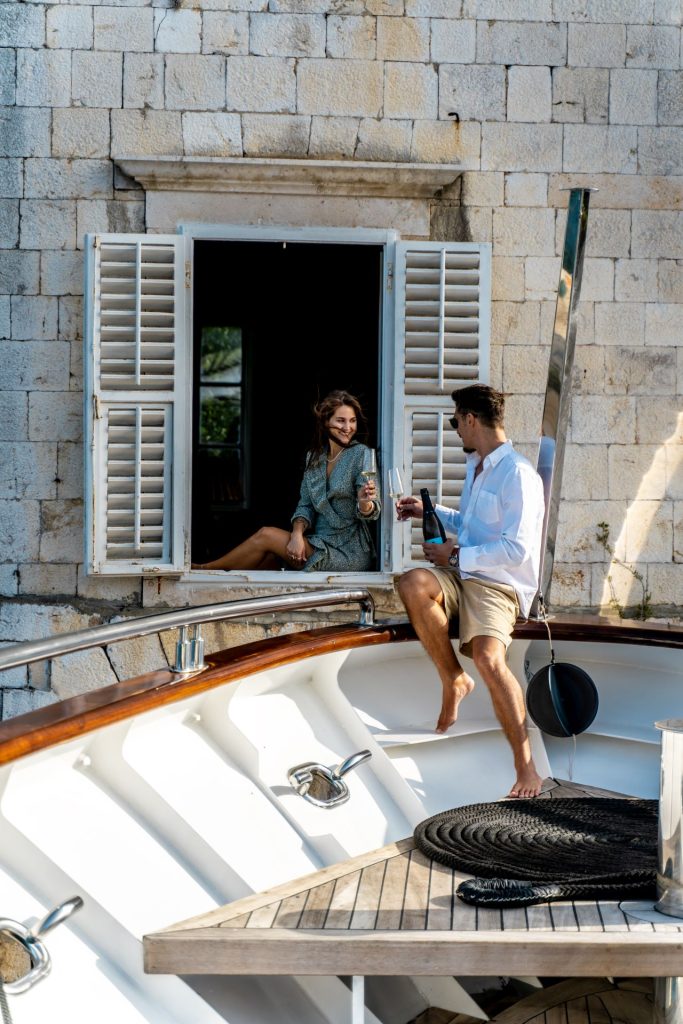 A woman sits in an open window of a stone building, smiling and holding a drink, while a man on a nearby boat reaches out to her with a glass, both appearing relaxed and enjoying the moment.