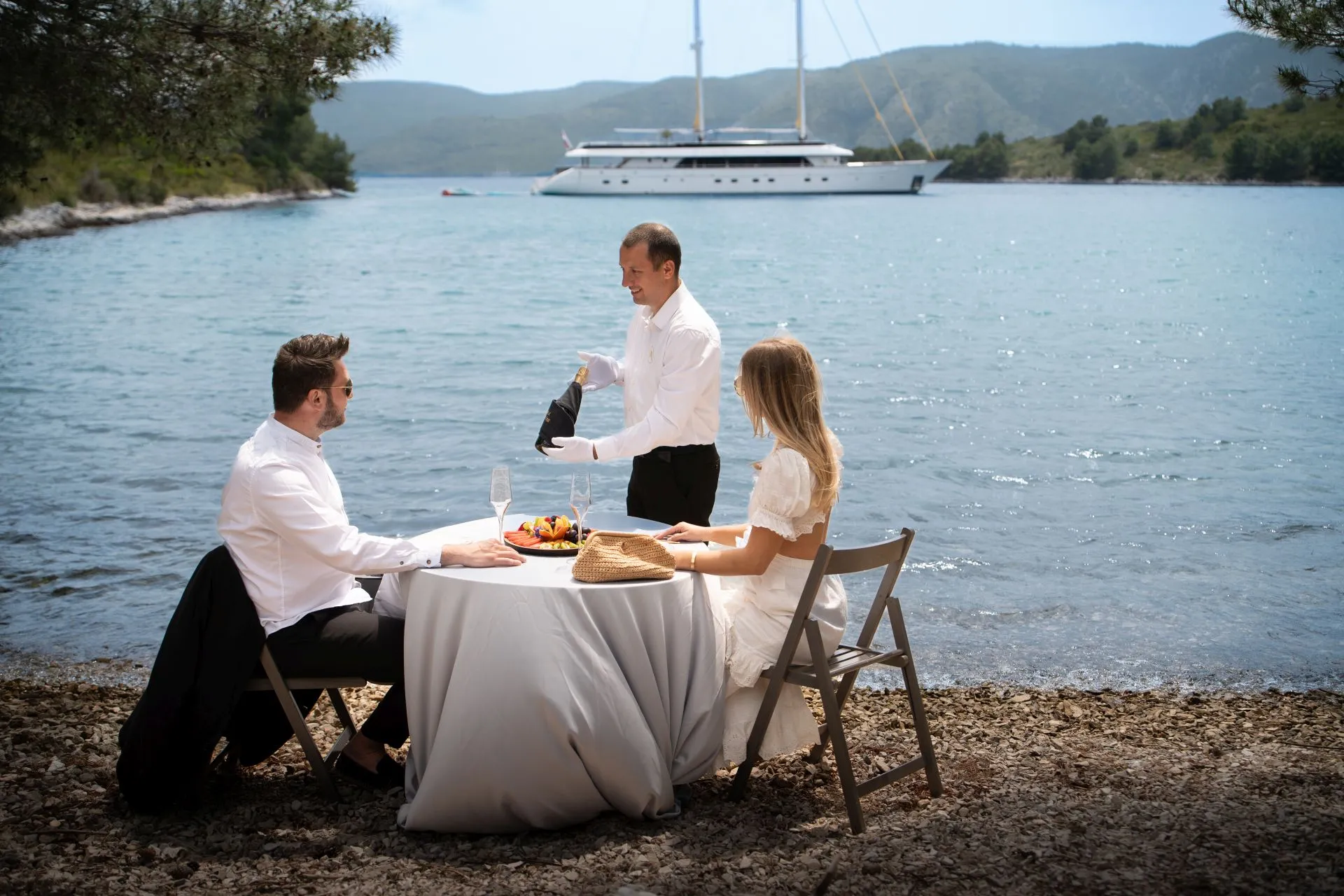 A waiter serves wine to a man and woman dining at a table by the water’s edge. A yacht is anchored in the background, and mountains rise beyond the calm sea under a clear sky.