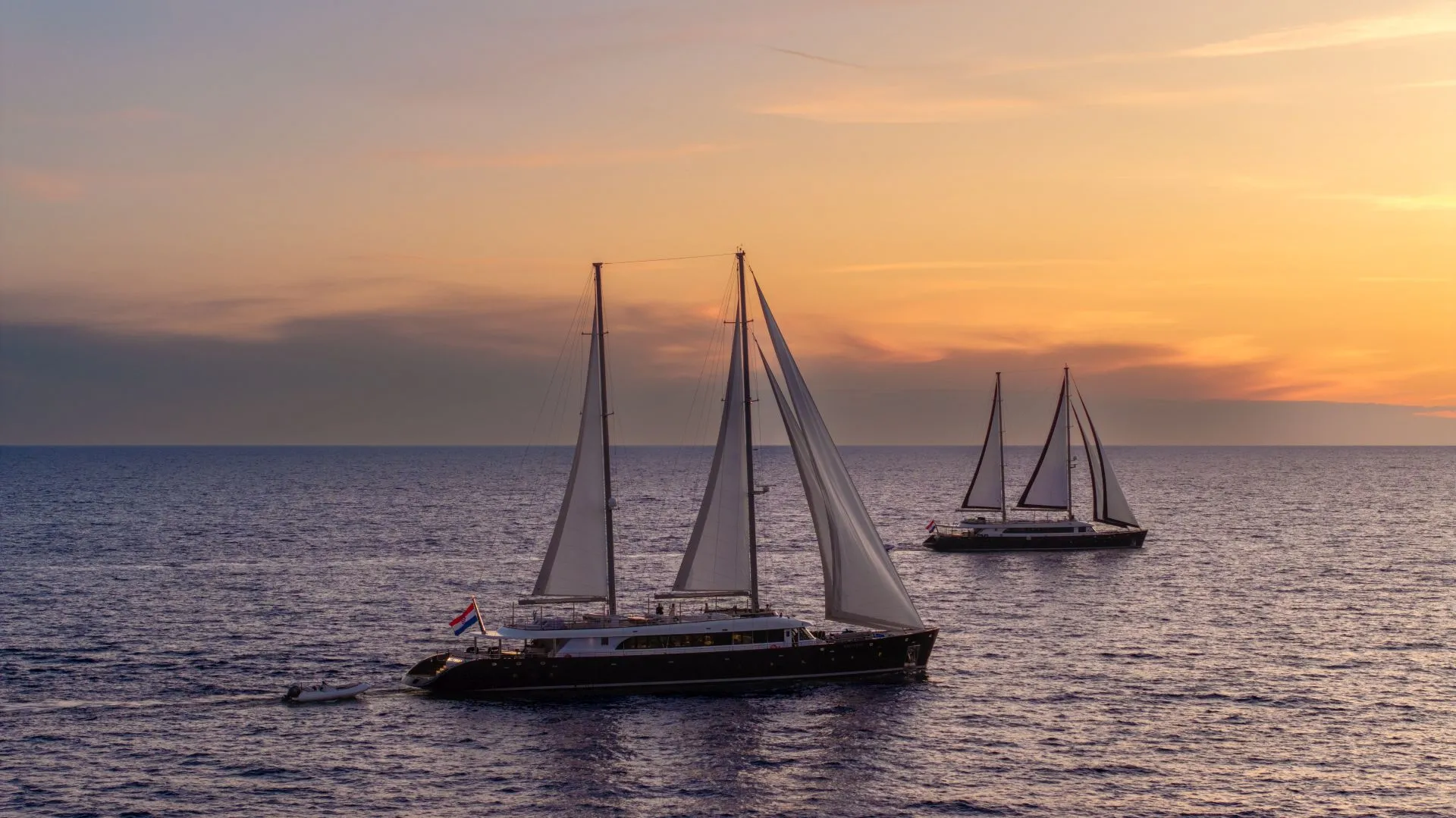 Two sailboats with white sails glide on calm water at sunset, with a colorful sky of orange, yellow, and purple hues in the background. One boat is closer to the foreground, the other farther away.