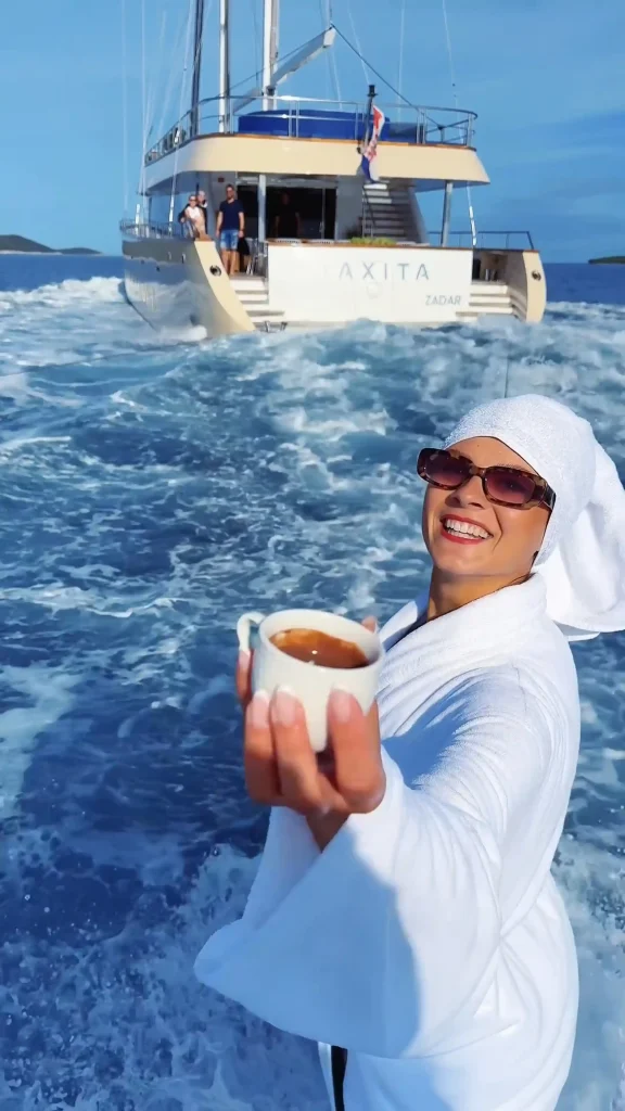 A smiling person in a white robe and towel on their head holds a cup of coffee toward the camera while standing on a boat, with a larger yacht and ocean waves in the background.
