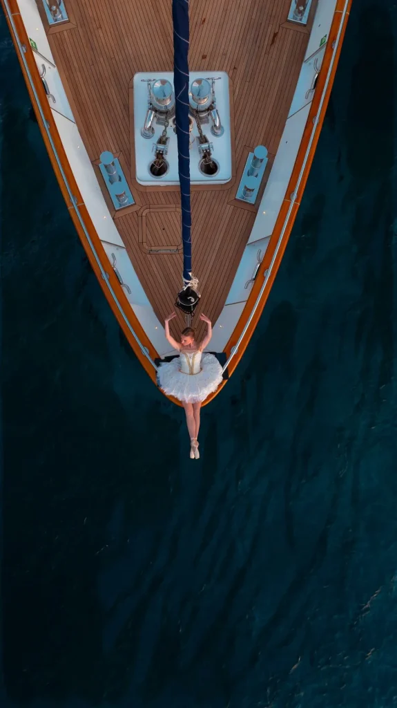 A ballerina in a white tutu lies on the bow of a yacht, her arms raised above her head, surrounded by deep blue water. The photo is taken from above, highlighting the contrast between the yacht and the sea.