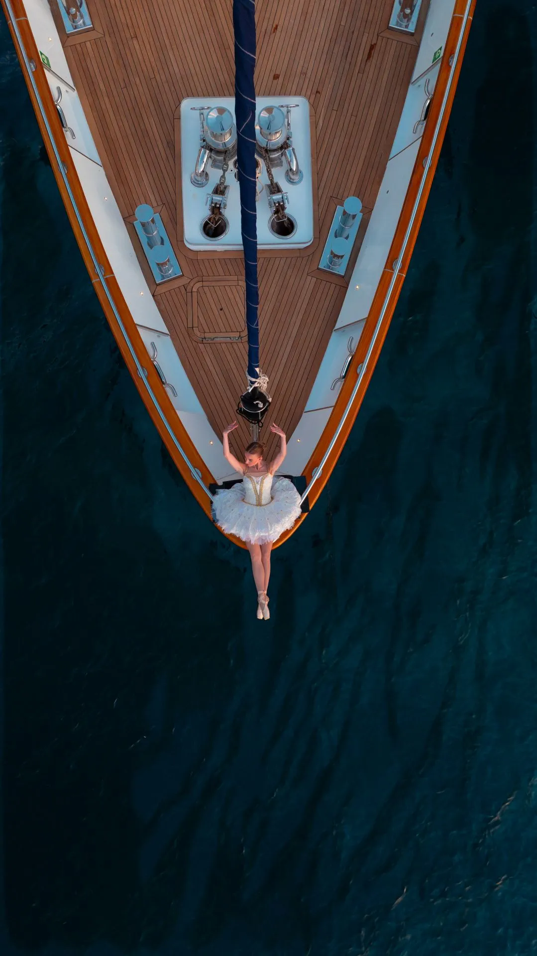 A ballerina in a white tutu lies on the bow of a yacht, her arms raised above her head, surrounded by deep blue water. The photo is taken from above, highlighting the contrast between the yacht and the sea.
