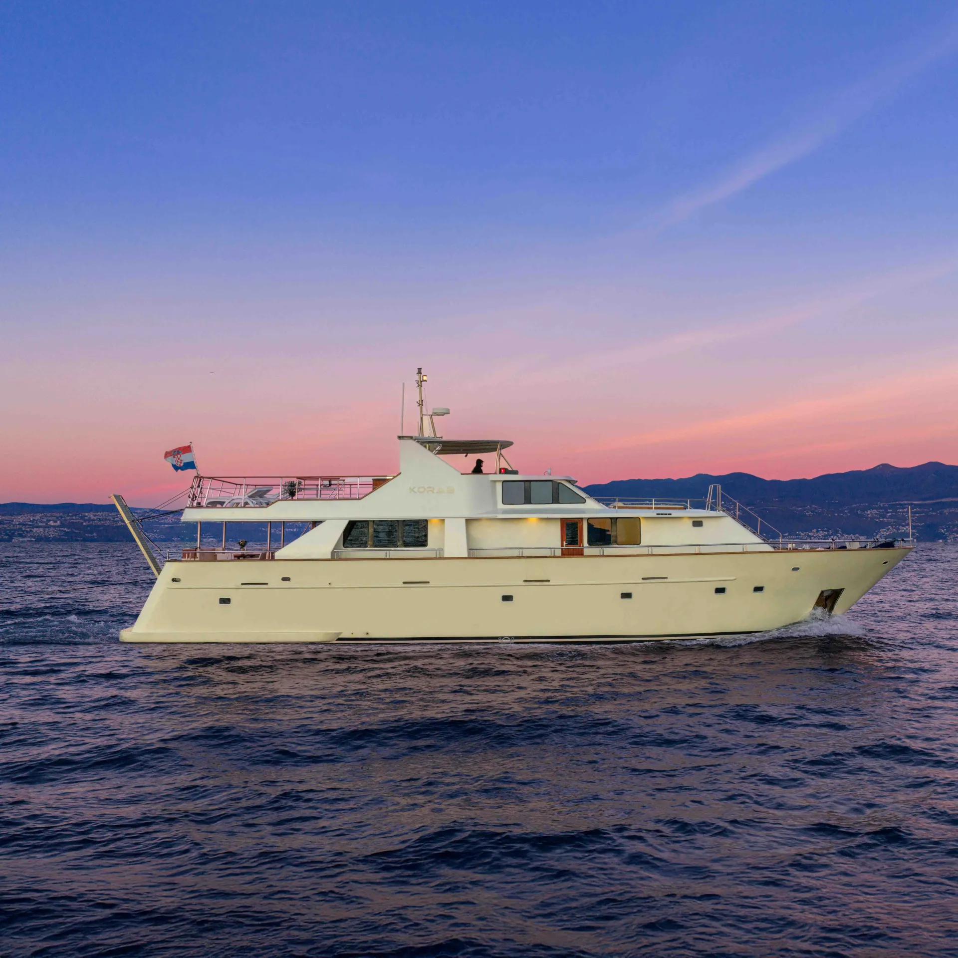 A cream-colored yacht cruises on calm water at sunset, with mountains and a pastel pink and blue sky in the background. The yacht has a flag at the stern and soft evening light reflects off the water.