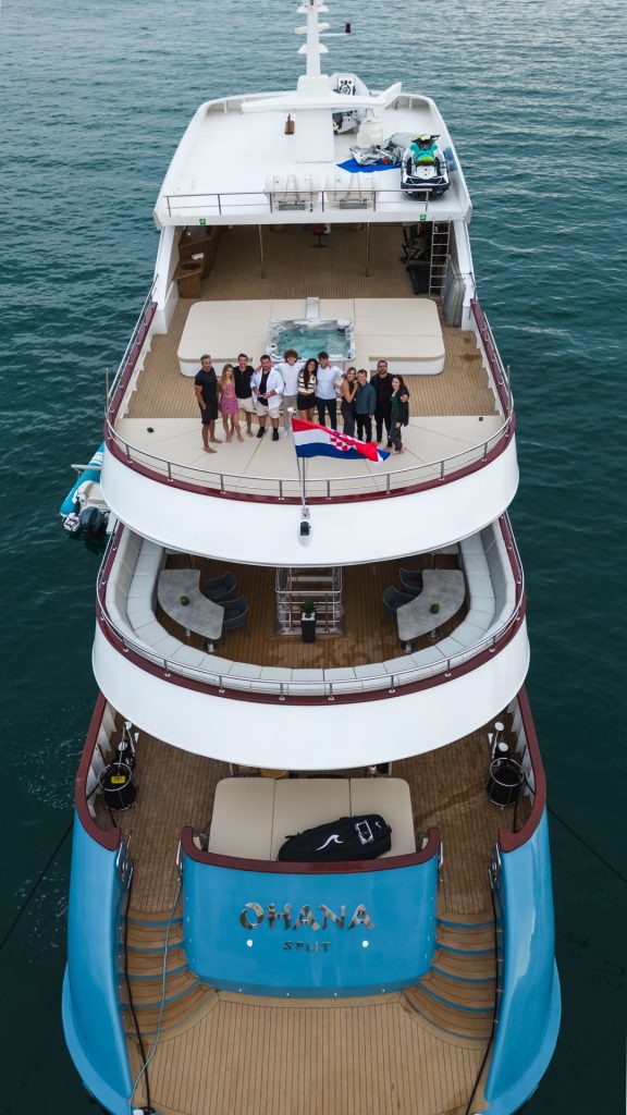 Aerial view of a large yacht named “Ohana Split” on the water, with a group of people standing together on the upper deck, smiling and posing for a photo behind a flag.