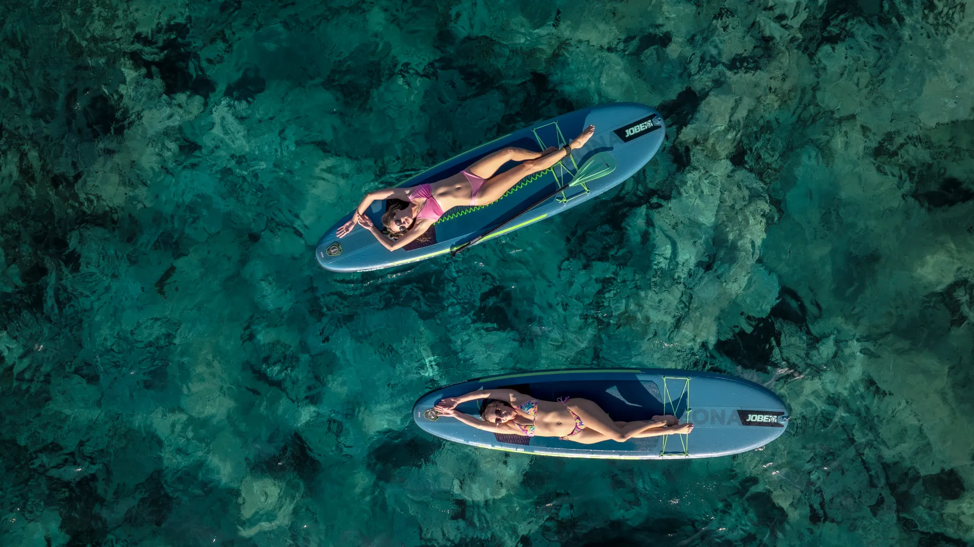 Aerial view of two people relaxing on paddleboards in clear, turquoise water. Both are lying on their backs, enjoying the sun, with the rocky seabed visible beneath them.