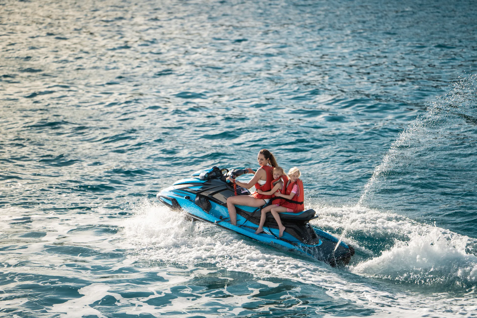 A woman and two young children, all wearing life jackets, ride together on a blue jet ski across the ocean, creating a spray of water as they speed through the waves.